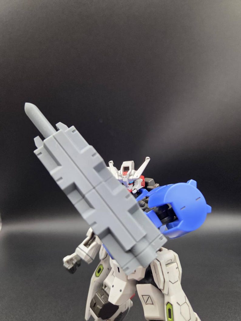 Op de afbeelding: Een wit, blauw en grijs gearticuleerd robotactiefiguur dat een groot, rechthoekig grijs wapen vasthoudt. Het figuur heeft een gedetailleerd hoofd met rode accenten, een blauw schouderstuk en mechanische beencomponenten met groene accenten.