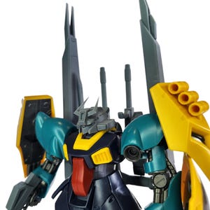 May include: A detailed model of a Gundam robot, featuring teal, yellow, gray, red, and navy blue components. The robot has large shoulder armor, multiple weapon attachments, and a detailed head design. The model is set against a white background.