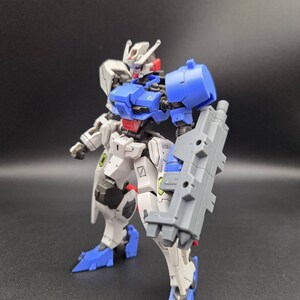 Op de afbeelding: Een beweegbaar robotactiefiguur met een wit, blauw en grijs kleurenschema. Het figuur heeft een gedetailleerd hoofd met antennes, een blauwe borstplaat, een rood middengedeelte en een groot grijs wapen aan de linkerarm. De benen zijn wit met blauwe voeten.