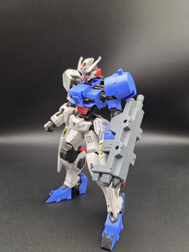 Op de afbeelding: Een beweegbaar robotactiefiguur met een wit, blauw en grijs kleurenschema. Het figuur heeft een gedetailleerd hoofd met antennes, een blauwe borstplaat, een rood middengedeelte en een groot grijs wapen aan de linkerarm. De benen zijn wit met blauwe voeten.