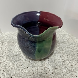 May include: A ceramic pitcher with a unique, wavy rim. The pitcher is decorated with a colorful glaze in shades of purple, red, green, and blue.