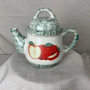 May include: A white ceramic teapot with a green speckled glaze and a lid. The teapot features a painted design of red apples with green leaves.