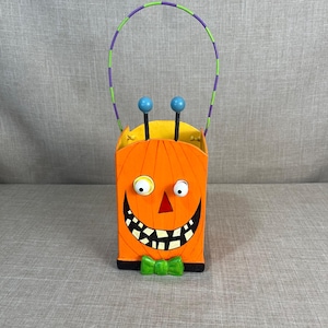 May include: A Halloween treat bucket shaped like a pumpkin with a big smile, googly eyes, and a green bow tie. The bucket has a purple and green handle.