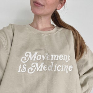 Op de afbeelding: Een beige sweatshirt met de tekst "Movement is Medicine" in witte cursieve letters.