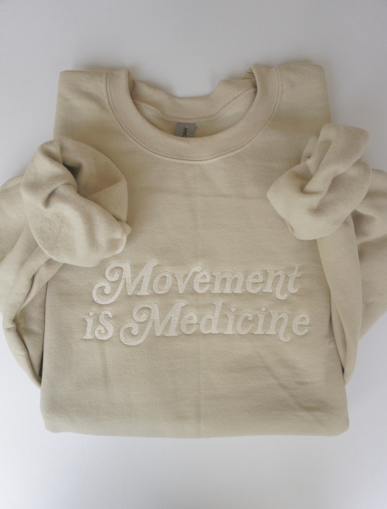 Op de afbeelding: Een beige sweatshirt met de tekst "Movement is Medicine" geborduurd in wit.