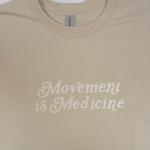 Op de afbeelding: Een beige T-shirt met de tekst "Movement is Medicine" in wit.