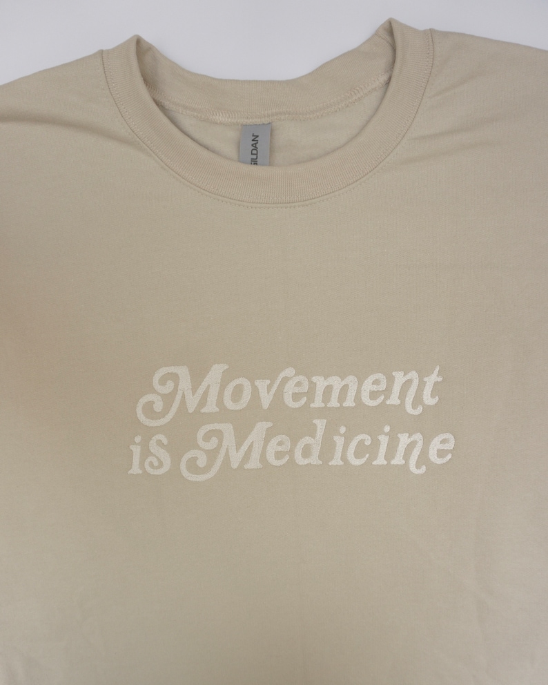 Op de afbeelding: Een beige T-shirt met de tekst "Movement is Medicine" in wit.