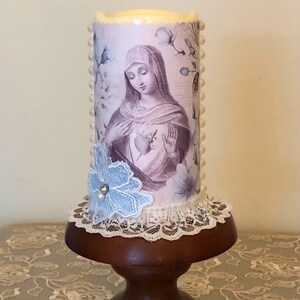 May include: A decorative candle featuring an image of a religious figure, adorned with a blue lace flower and white pearls. The candle sits on a dark wooden pedestal with a lace trim. The candle has a light purple floral design.