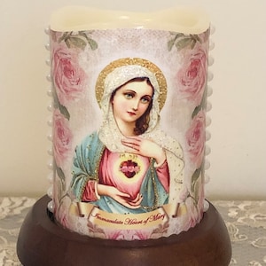 May include: A white pillar candle with a religious image of the Immaculate Heart of Mary. The candle is decorated with a pink rose pattern and white pearl beads around the edges. The candle is sitting on a wooden base.