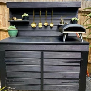 May include: Black outdoor kitchen with a wood-fired pizza oven, two storage cupboards, and a shelf with gold utensils hanging on hooks.
