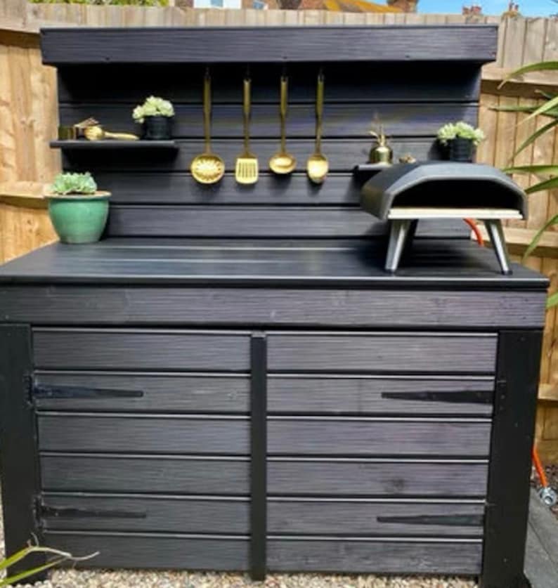 May include: Black outdoor kitchen with a wood-fired pizza oven, two storage cupboards, and a shelf with gold utensils hanging on hooks.