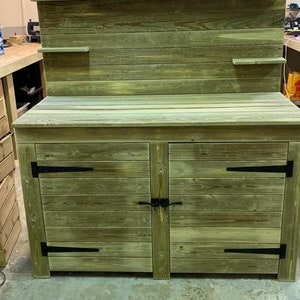 May include: A wooden cabinet with two doors and a top shelf. The cabinet is painted a light green colour and has black metal hinges. The cabinet is sitting on a concrete floor.