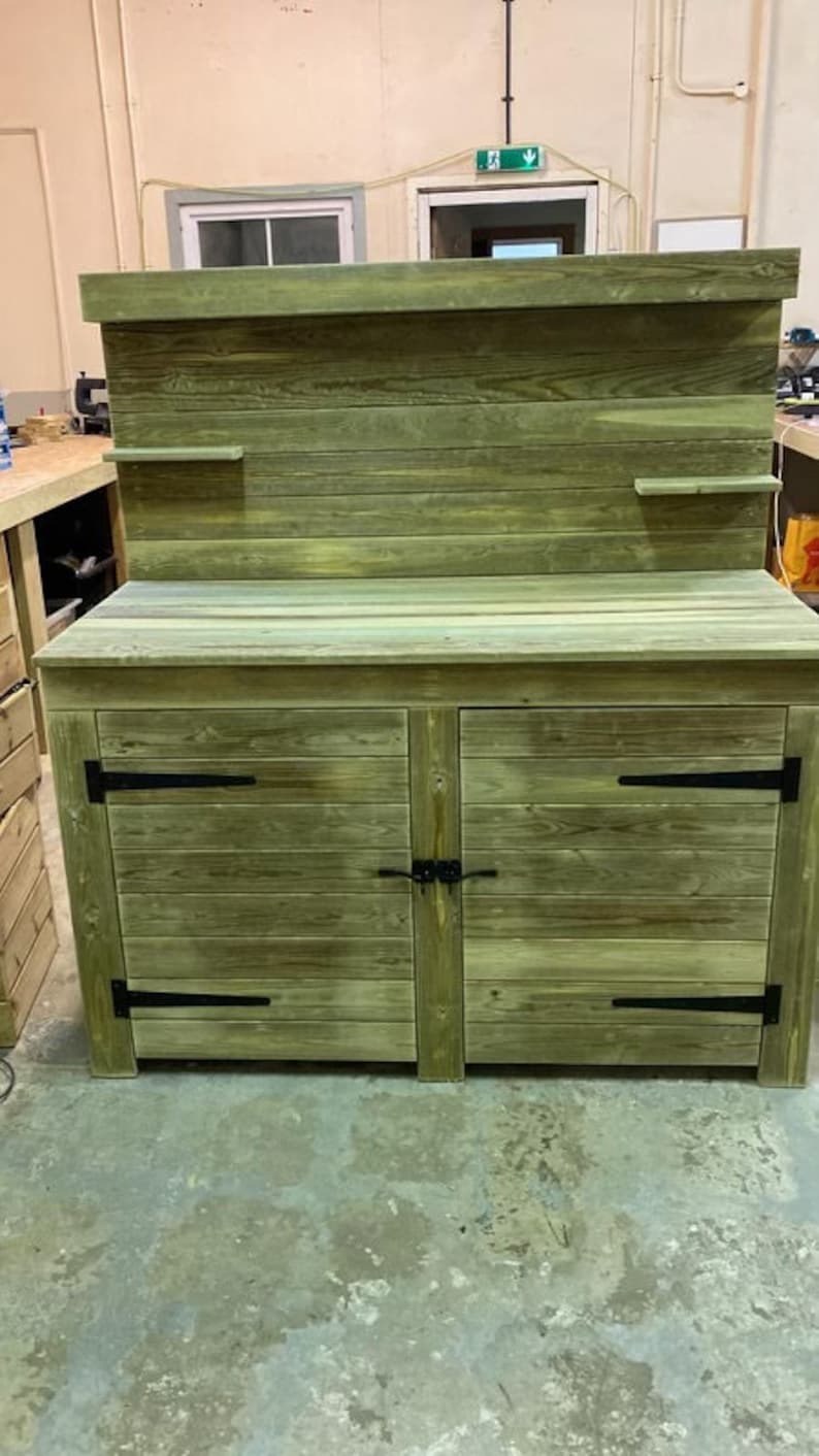 May include: A wooden cabinet with two doors and a top shelf. The cabinet is painted a light green colour and has black metal hinges. The cabinet is sitting on a concrete floor.