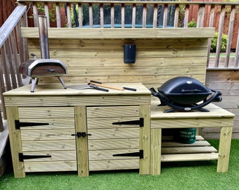 May include: A wooden outdoor kitchen with a pizza oven, grill, and storage. The structure is made of light-coloured wood and features a black grill, a pizza oven, and a black speaker. Includes a small propane gas cylinder.
