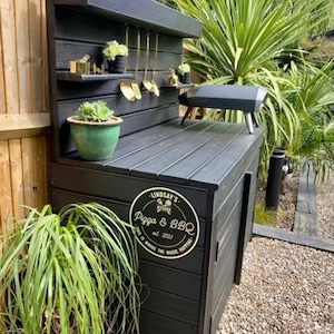 May include: Black outdoor kitchen counter with a wood-look finish. The counter has a built-in grill, storage cabinets, and a shelf for utensils. A gold sign with the text "Lindsay's Pizza & BBQ est. 2021" is attached to the counter.