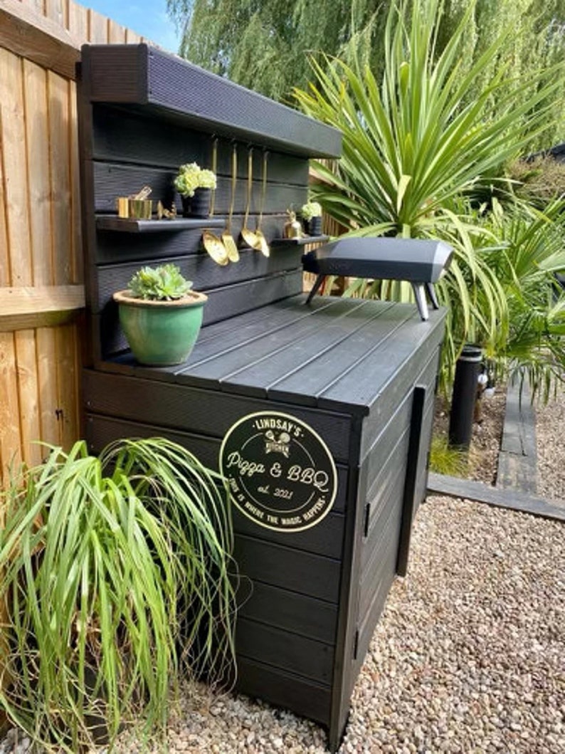May include: Black outdoor kitchen counter with a wood-look finish. The counter has a built-in grill, storage cabinets, and a shelf for utensils. A gold sign with the text "Lindsay's Pizza & BBQ est. 2021" is attached to the counter.