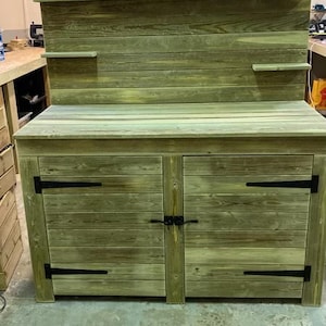 May include: A wooden cabinet with two doors and a top shelf. The cabinet is painted a light green colour and has black metal hinges. The cabinet is sitting on a concrete floor.