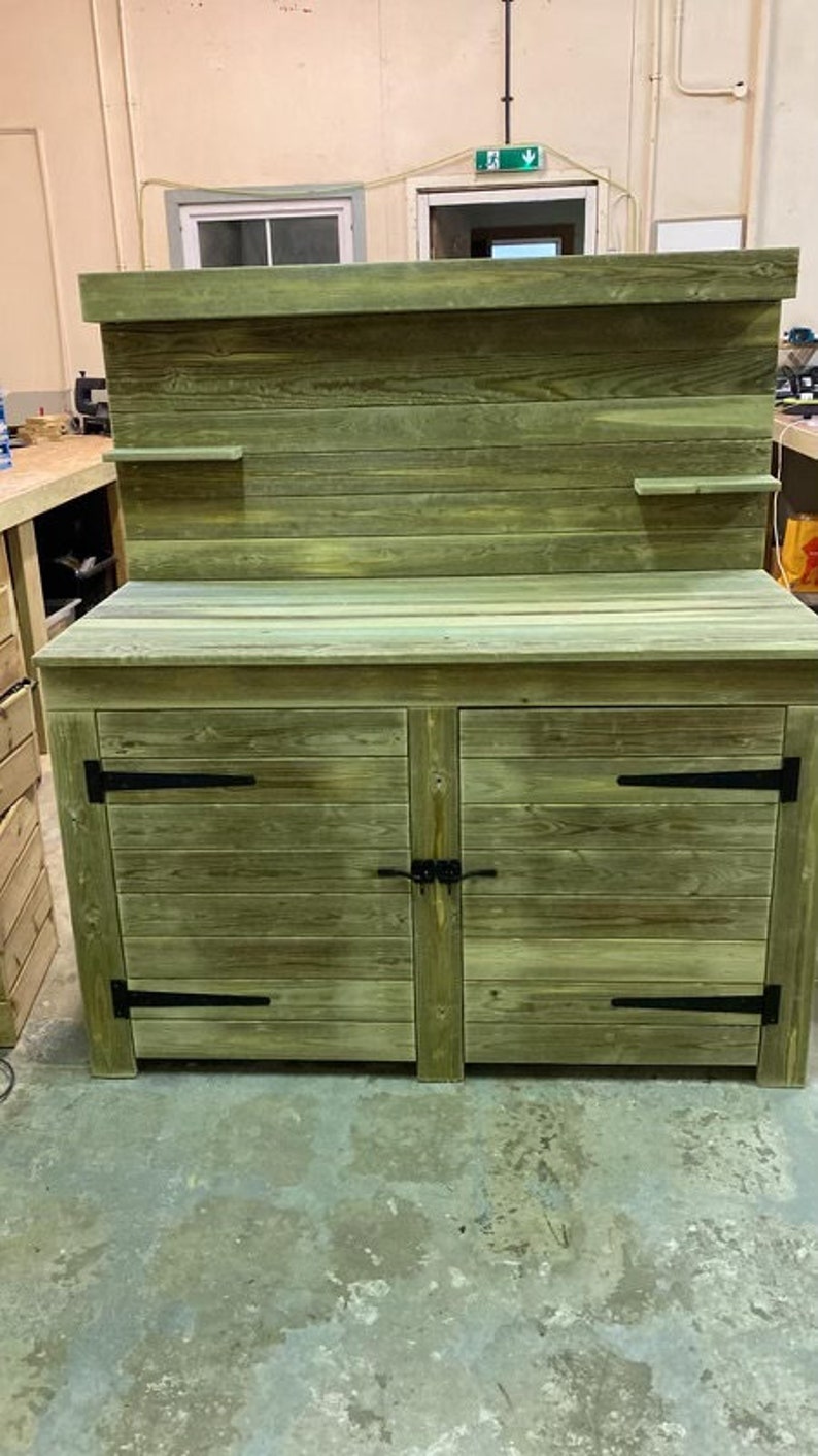 May include: A wooden cabinet with two doors and a top shelf. The cabinet is painted a light green colour and has black metal hinges. The cabinet is sitting on a concrete floor.