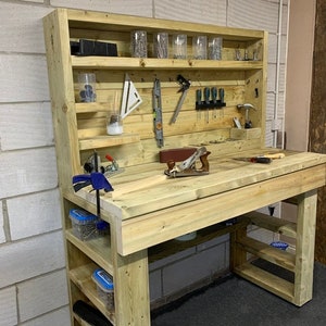 May include: A wooden workbench with a shelf unit above it. The workbench has a wooden top and a metal clamp. The shelf unit has several shelves and is filled with various tools and supplies. The workbench is made of unfinished wood and has a natural wood colour.