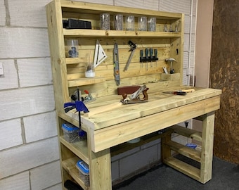 May include: A wooden workbench with a shelf unit above it. The workbench has a wooden top and a metal clamp. The shelf unit has several shelves and is filled with various tools and supplies. The workbench is made of unfinished wood and has a natural wood color.