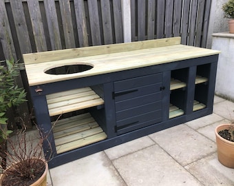 Puede incluir: Una gran estación de parrilla de madera para exteriores con un marco azul oscuro y una encimera de madera natural. Cuenta con un recorte circular para una parrilla, estantes y un armario con puerta. La unidad está diseñada para cocinar y entretener al aire libre.