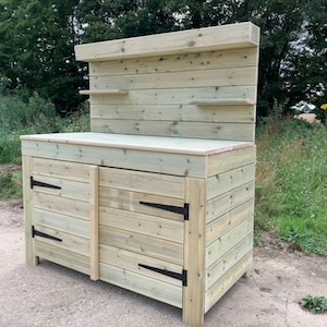 May include: A wooden garden work bench with two doors and a shelf. The bench is made of natural wood with a light brown finish. The bench has black metal hinges.