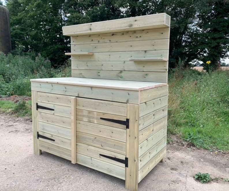 May include: A wooden garden work bench with two doors and a shelf. The bench is made of natural wood with a light brown finish. The bench has black metal hinges.
