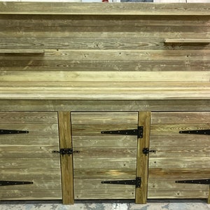 May include: A rustic wooden workbench with three doors and a top shelf. The workbench is made of reclaimed wood and has a natural finish. The doors have black metal hinges and latches.