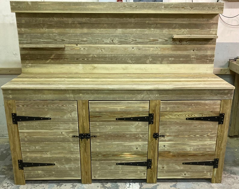May include: A rustic wooden workbench with three doors and a top shelf. The workbench is made of reclaimed wood and has a natural finish. The doors have black metal hinges and latches.