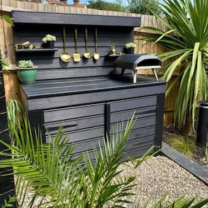 May include: A black outdoor kitchen with a wood-fired pizza oven, a wooden countertop, and a shelf with gold utensils. The kitchen is surrounded by plants and gravel.