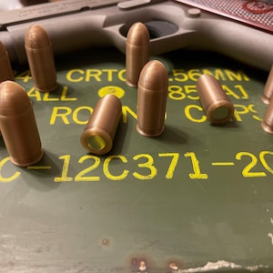 May include: A close-up of a green metal box with yellow text that reads "LC-12C371-204".  There are nine brown bullets on the box.  The bullets have a gold colored tip.