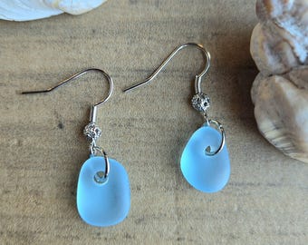 May include: A pair of light blue sea glass earrings with silver-colored hooks and decorative beads. The earrings have a smooth, rounded shape and a small hole at the top. The background includes seashells and a wooden surface.