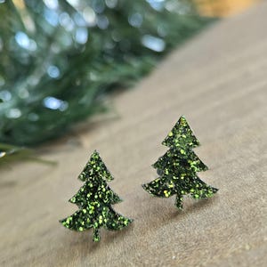 May include: Two green glitter Christmas tree stud earrings. The earrings are shaped like stylized evergreen trees with multiple layers of branches. The glitter is a mix of green shades, and the earrings are set against a blurred background.