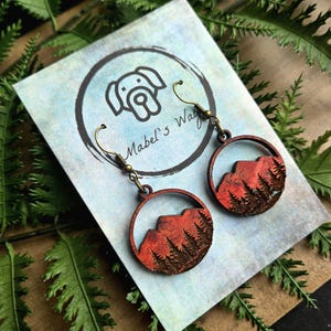 May include: Wooden dangle earrings featuring a mountain and forest design within a circular frame. The earrings have a red and brown color scheme, with brass-colored ear wires. The background includes a card with a dog logo and the text "Mabel's Way".