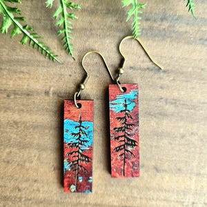 May include: Rectangular wooden earrings with a nature-inspired design. Each earring features a red background with a blue section and a black tree silhouette. The earrings have gold-colored ear wires.