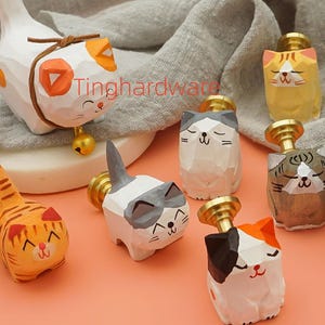 May include: A collection of hand-carved wooden cat figurines in various colors and patterns, including orange, white, gray, and black. Each cat has a brass-colored knob, suggesting they are drawer pulls or decorative accents.