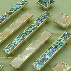 May include: A selection of cabinet hardware, including long pulls and square knobs. The pulls and knobs have a gold finish and are inlaid with iridescent abalone shell and cream-coloured material. The hardware is arranged on a light green surface.