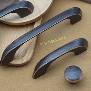 May include: Bronze-toned cabinet hardware set, including handles and a knob. The handles have a curved design and are displayed on a light brown surface. The text "Tinghardware" is visible.