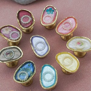 May include: A selection of decorative cabinet knobs with gold-coloured bases. Each knob features a unique design with colourful resin and embedded elements like flowers and pearls. The knobs are arranged on a pink surface.
