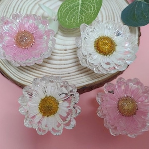 Könnte beinhalten: Vier klare, harzförmige Objekte in Blumenform, zwei rosa und zwei weiße, mit gelben Zentren. Die Blumen sind auf einer Holzscheibe und einem rosa Hintergrund angeordnet. Die Blütenblätter haben ein strukturiertes, kristallines Aussehen.