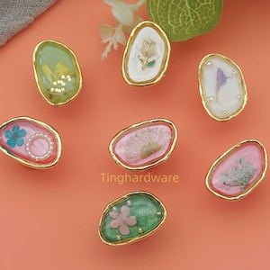 May include: Seven decorative resin knobs with gold-colored edges. Each knob features a unique design with dried flowers, pearls, and other embellishments in shades of pink, green, and white. The knobs are arranged on a peach-colored surface.