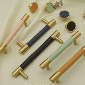 Può includere: Un set di ferramenta per mobili, tra cui maniglie e pomelli color ottone. Le maniglie presentano manici rivestiti in pelle colorata in rosa, blu navy, nero, verde menta e beige. I pomelli hanno centri colorati coordinati. La ferramenta è esposta su una superficie color crema.