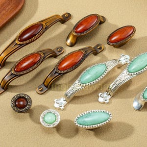 May include: A collection of decorative cabinet hardware, including handles and knobs. The handles feature a bronze finish with oval, reddish-brown center stones. Other pieces have silver finishes with green center stones. The hardware is designed for furniture.