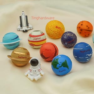 May include: A collection of colorful planet-shaped figurines, including the Sun, Earth, and Mars, along with a space shuttle and astronaut figures. The planets are painted with details and labeled with their names.
