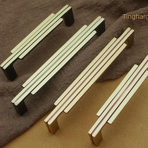May include: Four gold-toned cabinet pulls with a linear design and black accents. The pulls have a rectangular shape with three parallel lines and are arranged on a brown surface. The text "Tinghardware" is visible.