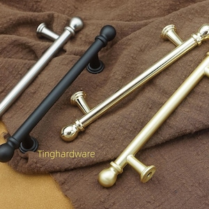 Può includere: Quattro maniglie per mobili in argento, nero e oro sono appoggiate su un tessuto marrone. Le maniglie hanno un design a barra con estremità arrotondate e montanti a forma di T. Il testo "Tinghardware" è visibile.