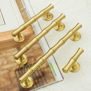 Puede incluir: Tiradores de armario dorados con estilo de bambú. El herraje tiene un asa cilíndrica con un diseño de tallo de bambú, montada sobre una base circular. Los tiradores están dispuestos sobre una revista con el texto "Tingharda".