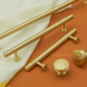 以下が含まれることがあります： ゴールドトーンのキャビネットハードウェアのコレクション。長いバープルと丸いノブが含まれています。 クリーム色の生地とオレンジ色の表面に表示されています。