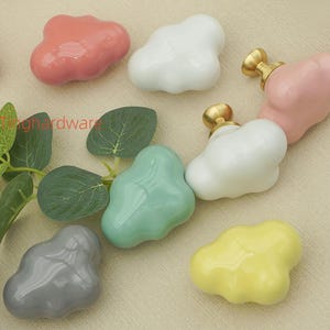 May include: A selection of ceramic cabinet knobs in a range of colours and cloud-like forms. Colours include pink, red, white, blue, grey, green, and yellow. Some knobs have gold-coloured hardware. Decorative furniture hardware.