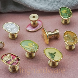 May include: A selection of decorative cabinet knobs with brass bases and oval resin tops. The resin tops incorporate floral elements and come in various colours, including green, yellow, and pink. These knobs are designed for furniture and home decor.